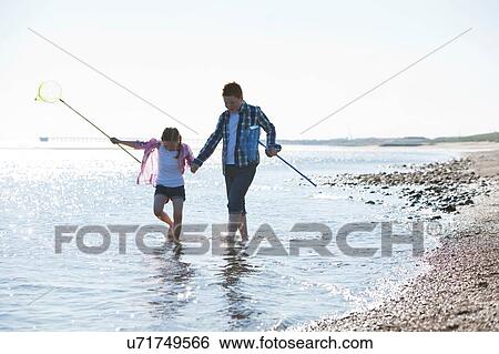 Stock Photograph - "Girl and boy paddling in water holding hands, carrying fishing nets, looking down". Fotosearch