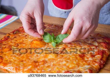 Stock Photography - Hands of woman placing basil on pizza at kitchen counter. Fotosearch