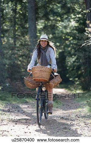 Stock Image - Mature woman cycling with foraging baskets on forest path. Fotosearch