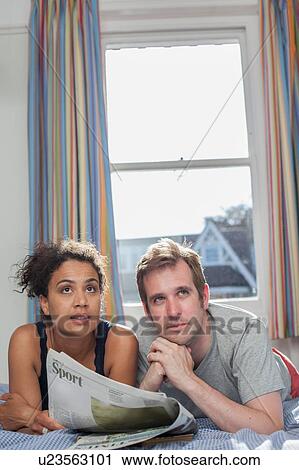 Stock Image - "Mid adult couple lying on bed, looking at newspaper". Fotosearch