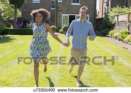 Stock Image - "Mid adult couple running, hand in hand, in garden". Fotosearch