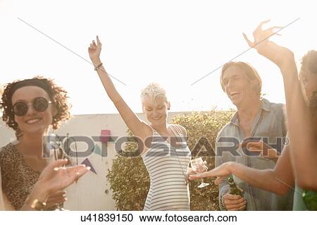 Stock Image - Mid adult man and young women drinking and dancing at poolside party. Fotosearch
