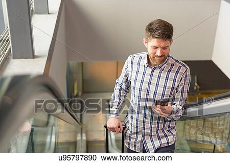 Stock Image - Mid adult man on esculator using smartphone looking down smiling. Fotosearch
