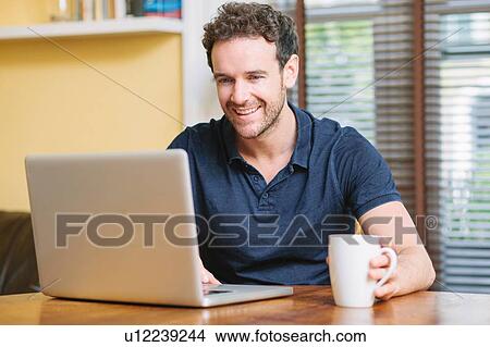 Mid adult man sitting infront of window holding cup using laptop smiling View Large Photo Image Picture - Mid adult man sitting infront of window holding cup using laptop smiling. Fotosearch