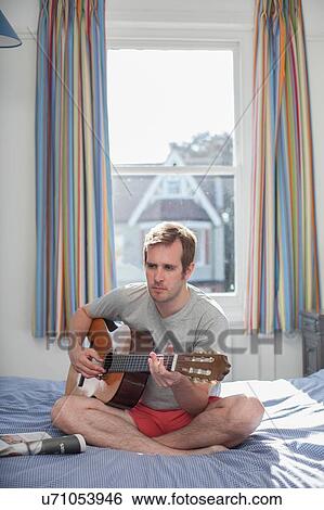 Stock Photograph - "Mid adult man sitting on bed, playing guitar". Fotosearch