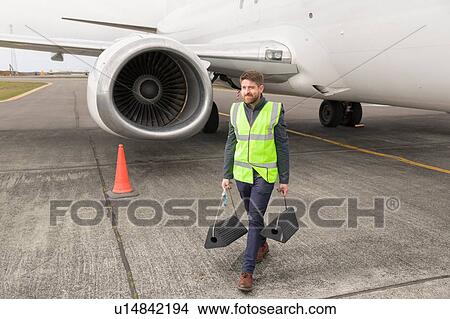 Picture - Mid adult man wearing high visability jacket walking past airplane engine carrying chocks. Fotosearch