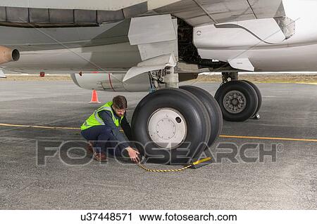 Stock Image - Mid adult man wearing high visability jacket kneeling next to airplane wheels placing chock. Fotosearch
