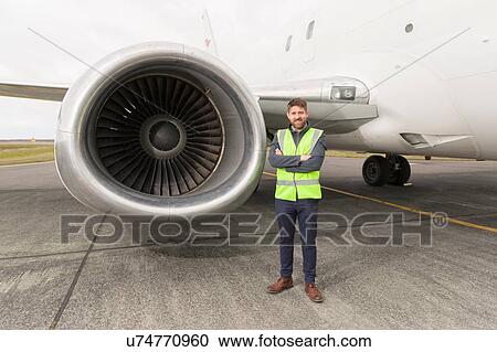 Stock Image - "Mid adult man wearing high visability jacket standing next to airplane engine, arms folded, looking at camera". Fotosearch