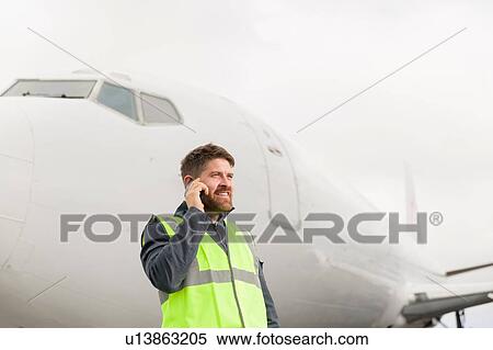 "Mid adult man wearing high visability jacket standing in front of airplane using smartphone, looking away smiling" View Large Photo Image Stock Photography - "Mid adult man wearing high visability jacket standing in front of airplane using smartphone, looking away smiling". Fotosearch