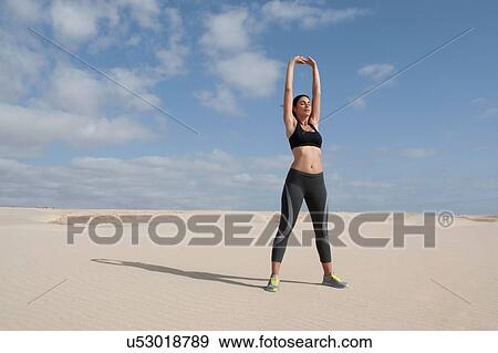 Stock Photo - Mid adult woman exercising with arms raised on beach. Fotosearch