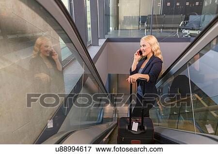 "Mid adult woman holding suitcase on esculator using smartphone, looking away smiling" View Large Photo Image Stock Photo - "Mid adult woman holding suitcase on esculator using smartphone, looking away smiling". Fotosearch