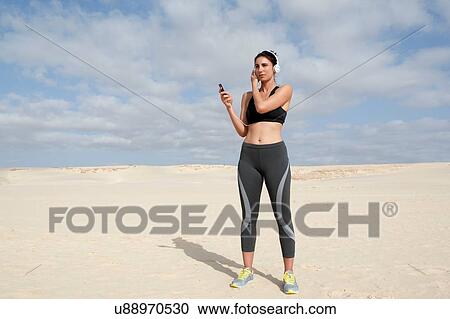 Stock Image - Mid adult woman selecting headphone music whilst exercising at beach. Fotosearch