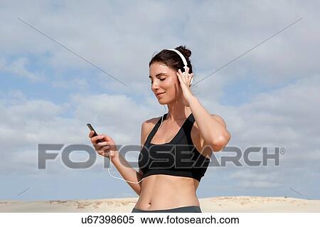 Stock Photography - Mid adult woman selecting music for headphones whilst exercising at beach. Fotosearch