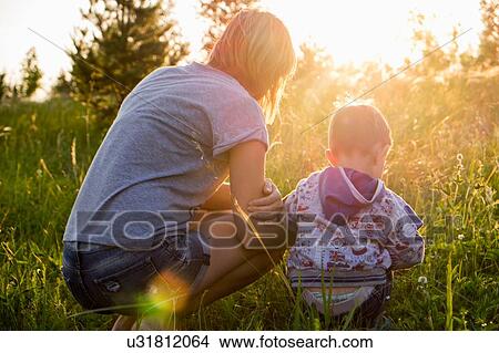 Picture - "Mother and son, crouching in field at sunset, rear view". Fotosearch