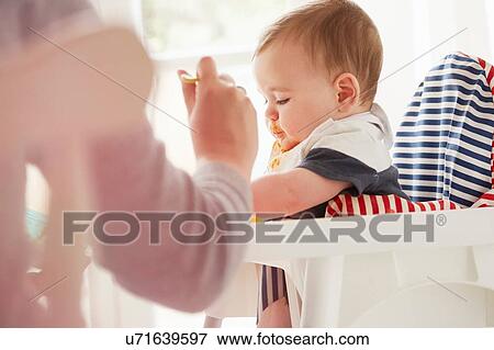 Stock Photo - Mother feeding baby boy in baby chair. Fotosearch
