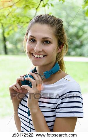 Stock Image - Portrait of young woman holding headphones looking at camera smiling. Fotosearch