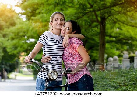 Stock Image - "Portrait of young women with bicycle, hugging, looking at camera smiling". Fotosearch