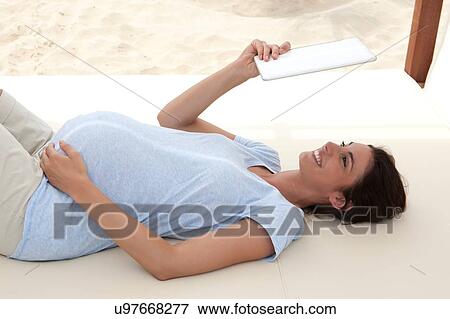 Stock Photo - Pregnant mid adult woman lying on beach daybed looking up at digital tablet. Fotosearch