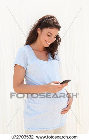 Stock Image - Pregnant mid adult woman reading texts on smartphone. Fotosearch