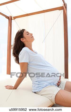 Stock Image - Pregnant mid adult woman sitting on daybed with hand on stomach. Fotosearch