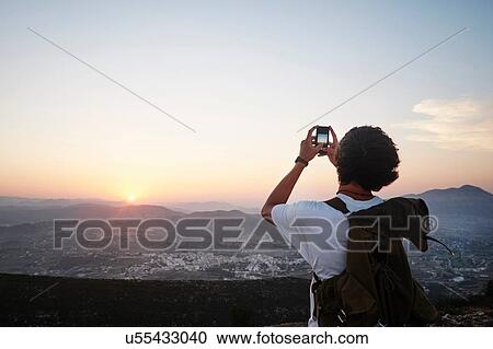 Stock Image - "Rear view of young man photographing landscape and sunset on smartphone, Javea, Spain". Fotosearch