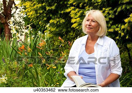 Stock Image - Senior woman sitting in garden holding book. Fotosearch