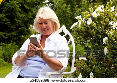 Senior woman sitting in garden looking at smartphone View Large Photo Image Stock Photo - Senior woman sitting in garden looking at smartphone. Fotosearch