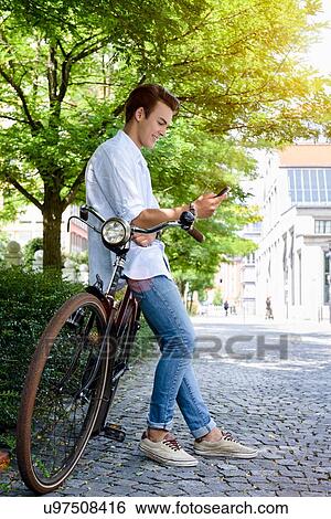 Stock Photograph - Side view of young man leaning against bicycle looking at smartphone smiling. Fotosearch