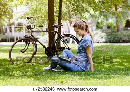 Side view of young woman sitting cross legged on grass next to bicycle using digital tablet View Large Photo Image Stock Image - Side view of young woman sitting cross legged on grass next to bicycle using digital tablet. Fotosearch
