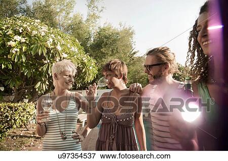 "Small group of friends standing holding drinks talking and smiling, lens flare" View Large Photo Image Stock Photo - "Small group of friends standing holding drinks talking and smiling, lens flare". Fotosearch