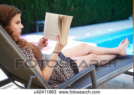 Stock Image - Teenage girl reading book on lounger at swimming pool. Fotosearch