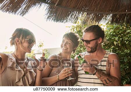 Stock Photo - "Three friends waist up standing under straw parasol holding drinks, smiling". Fotosearch