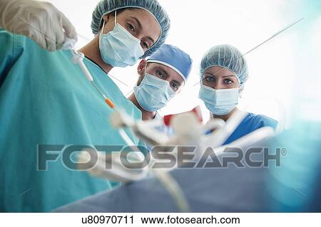Stock Image - Three surgeons concentrating during operation in hospital operating theatre. Fotosearch