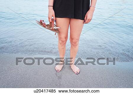Stock Photography - Waist down of young woman on beach holding sandals. Fotosearch