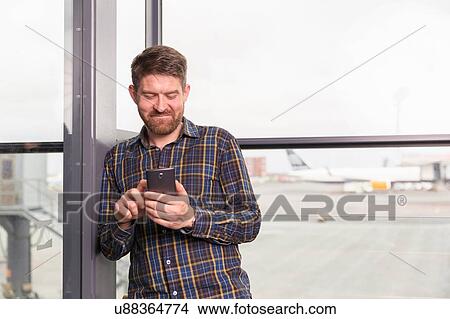 Waist up front view of mid adult man standing in front of airport window using smartphone View Large Photo Image Picture - Waist up front view of mid adult man standing in front of airport window using smartphone. Fotosearch