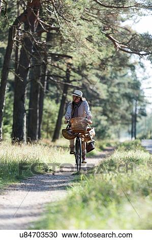 Stock Image - Woman cycling on forest path with foraging baskets. Fotosearch