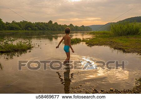 Stock Photo - "Young boy paddling in lake at sunset, rear view". Fotosearch