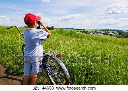 Stock Photo - "Young boy standing with bicycle beside field, looking at landscape through binoculars, rear view". Fotosearch