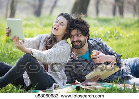 Stock Image - Young couple on picnic blanket taking digital tablet selfie in woodland. Fotosearch