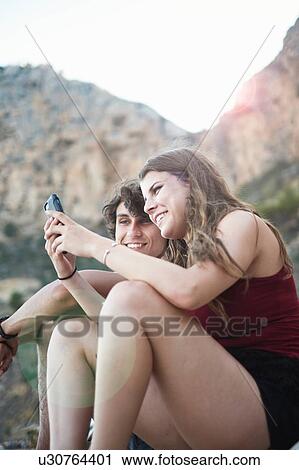 Stock Image - "Young man and teenage sister reading smartphone texts on rocky beach, Javea, Spain". Fotosearch