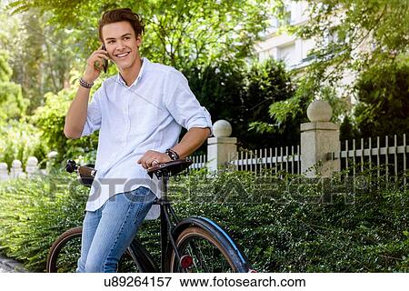 Stock Photo - "Young man leaning against bicycle using smartphone, looking away smiling". Fotosearch