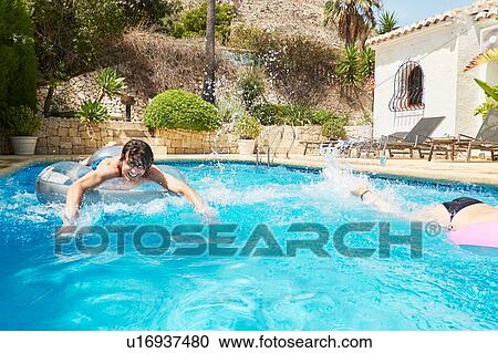 Stock Image - Young man racing teenage sister on inflatables in swimming pool. Fotosearch