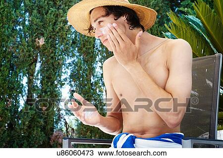 Stock Image - Young man wearing sunhat applying suncream to face. Fotosearch
