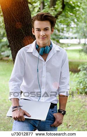 "Young man with headphones leaning against tree, hand in pocket, holding laptop looking at camera smiling" View Large Photo Image Stock Photo - "Young man with headphones leaning against tree, hand in pocket, holding laptop looking at camera smiling". Fotosearch