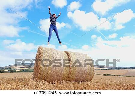 Stock Photography - Young woman jumping on top of haystacks in harvested field. Fotosearch