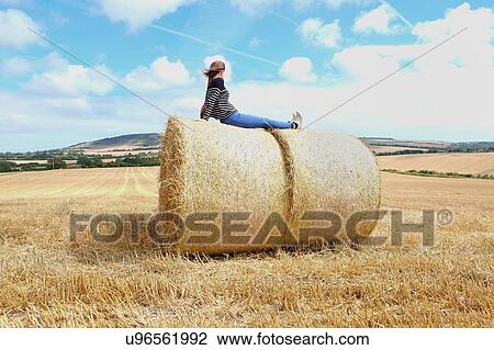 Stock Image - Young woman looking out from top of haystacks in harvested field. Fotosearch