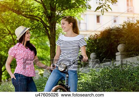 Stock Photograph - "Young woman on bicycle talking to young woman wearing panama hat, face to face smiling". Fotosearch