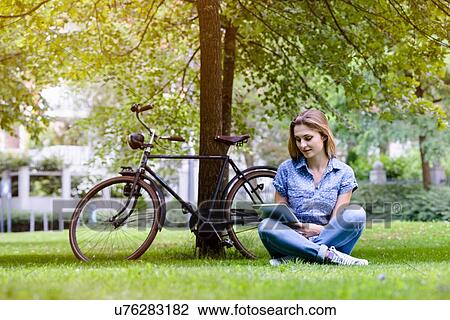 Young woman sitting cross legged on grass next to bicycle using digital tablet View Large Photo Image Stock Image - Young woman sitting cross legged on grass next to bicycle using digital tablet. Fotosearch