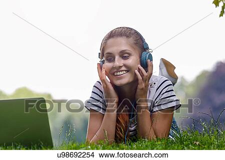 Young woman wearing headphones lying on grass resting on elbows looking at laptop smiling View Large Photo Image Picture - Young woman wearing headphones lying on grass resting on elbows looking at laptop smiling. Fotosearch
