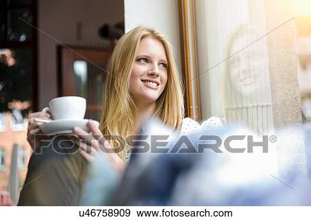 Stock Photo - "Young woman with long blond hair sitting holding coffee cup, looking away smiling". Fotosearch